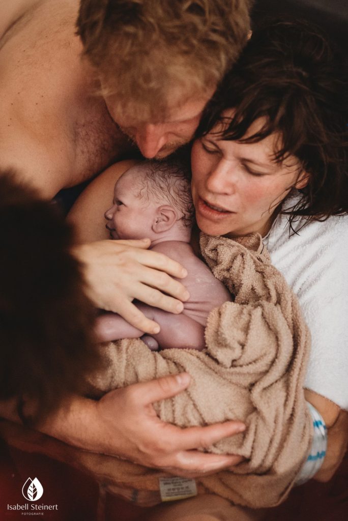 Birth photography showing women with newly born infant with a blanket loosely placed around him/her and man embracing them. 